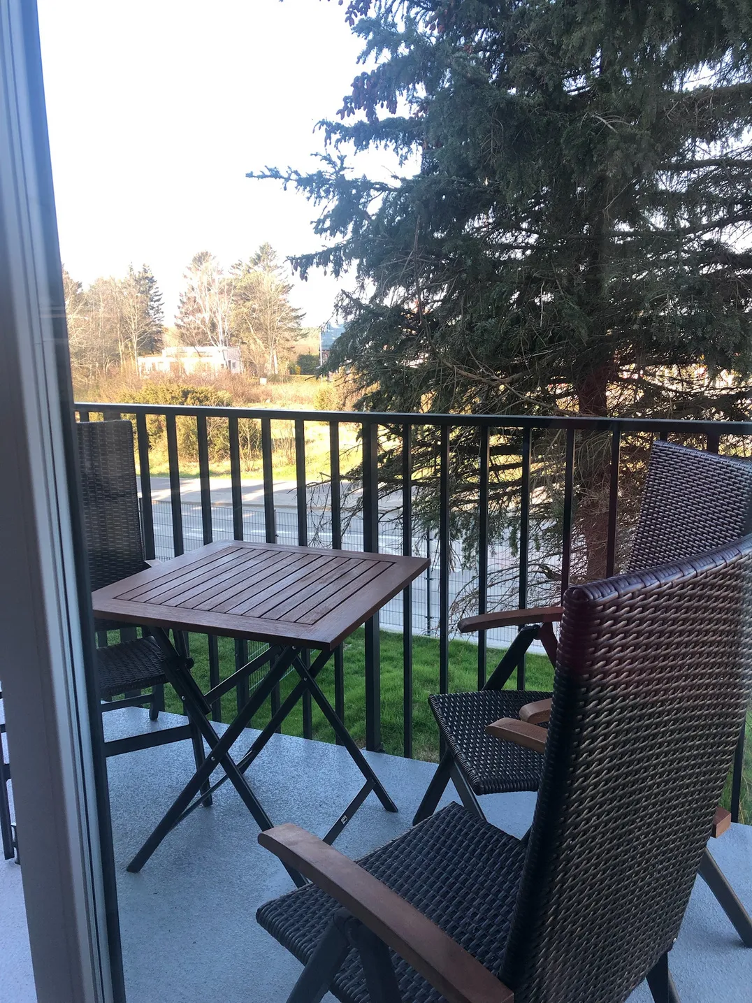 Balkon z drewnianym stolikiem i wiklinowymi fotelami, w tle sosny i trawnik