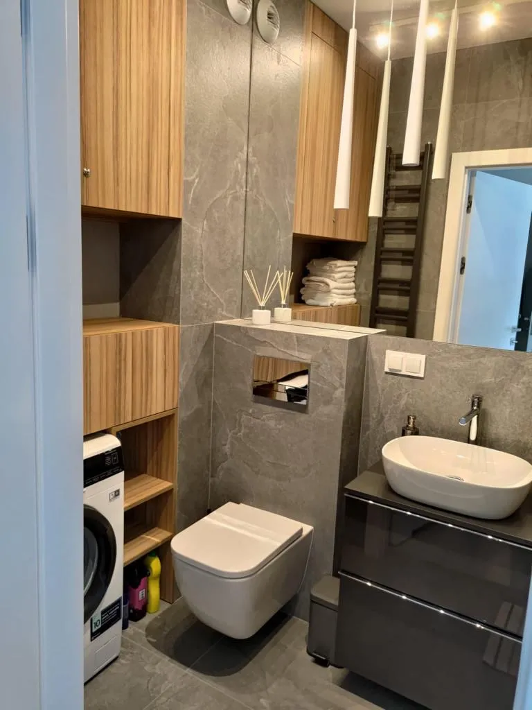 Łazienka z szarymi kamiennymi płytkami, drewnem, białą umywalką i designerskimi lampami